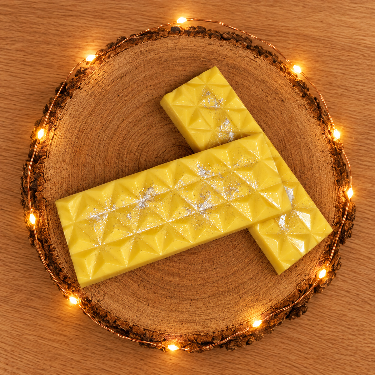 Vue de dessus de deux tablettes de fondant parfumé Fleur de Coton jaunes, disposées en croix sur un rondin de bois illuminé.