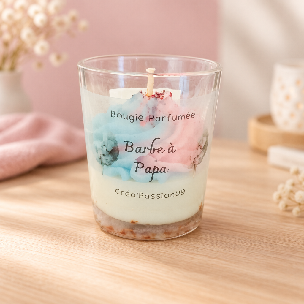 Bougie parfumée Barbe à Papa Créa'Passion09 en pot de verre, posée sur du bois clair devant un mur rose poudré